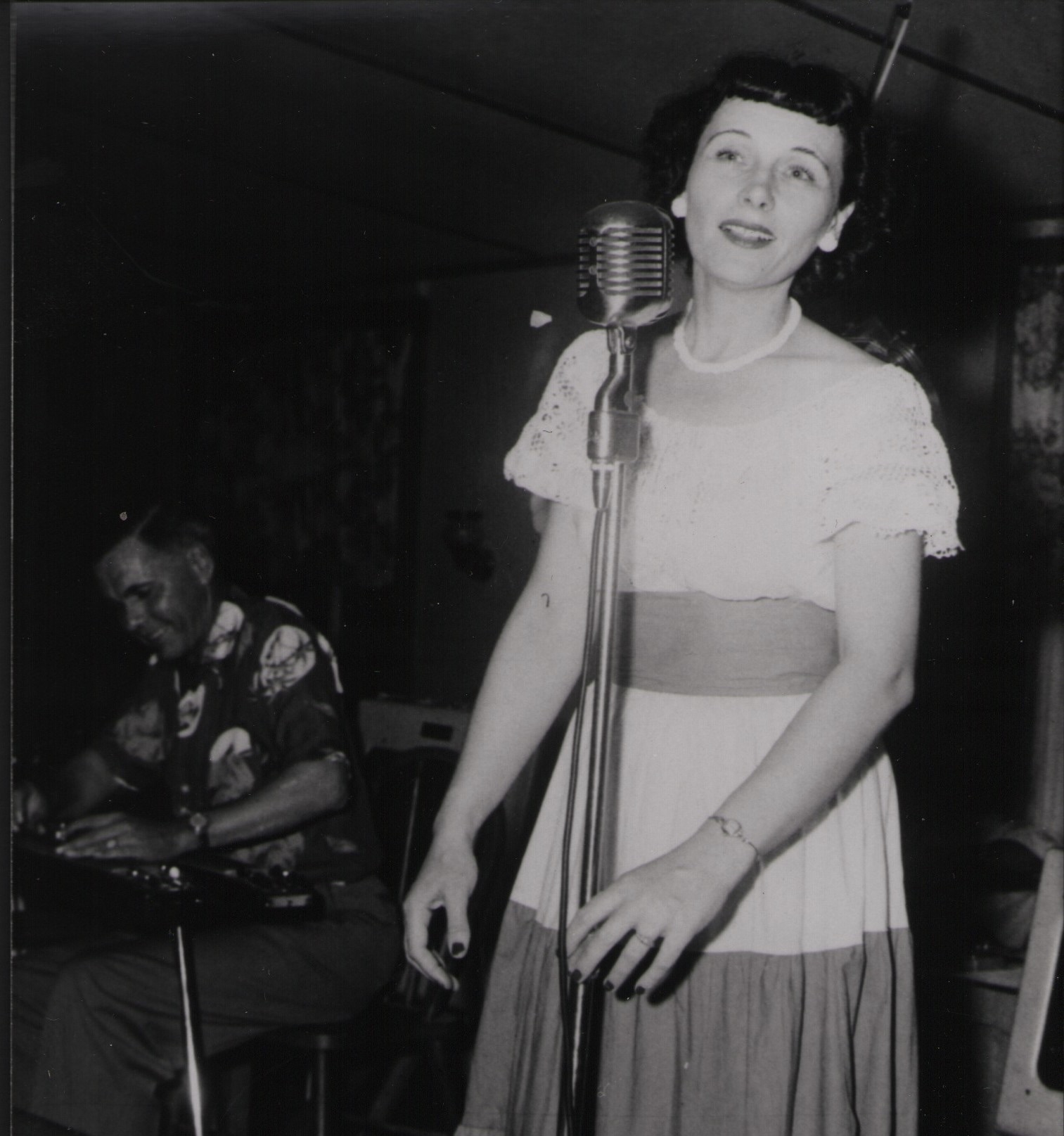 Dolores Fariss singing into a vintage microphone, wearing a tiered dress with lace detailing.