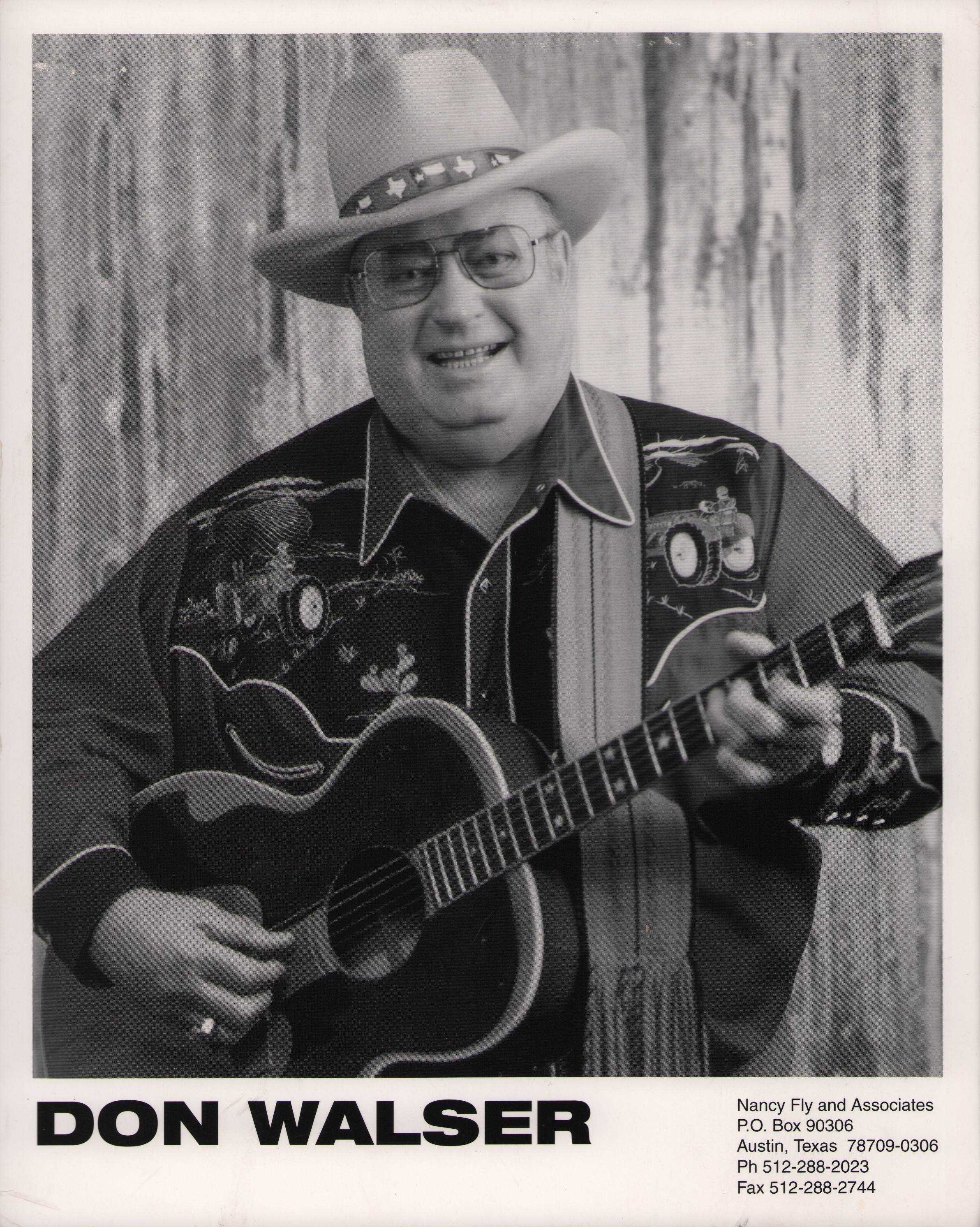 Don Walser holding an acoustic guitar and smiling. He is wearing a cowboy hat, glasses, and a Western-style shirt with embroidered tractor designs.