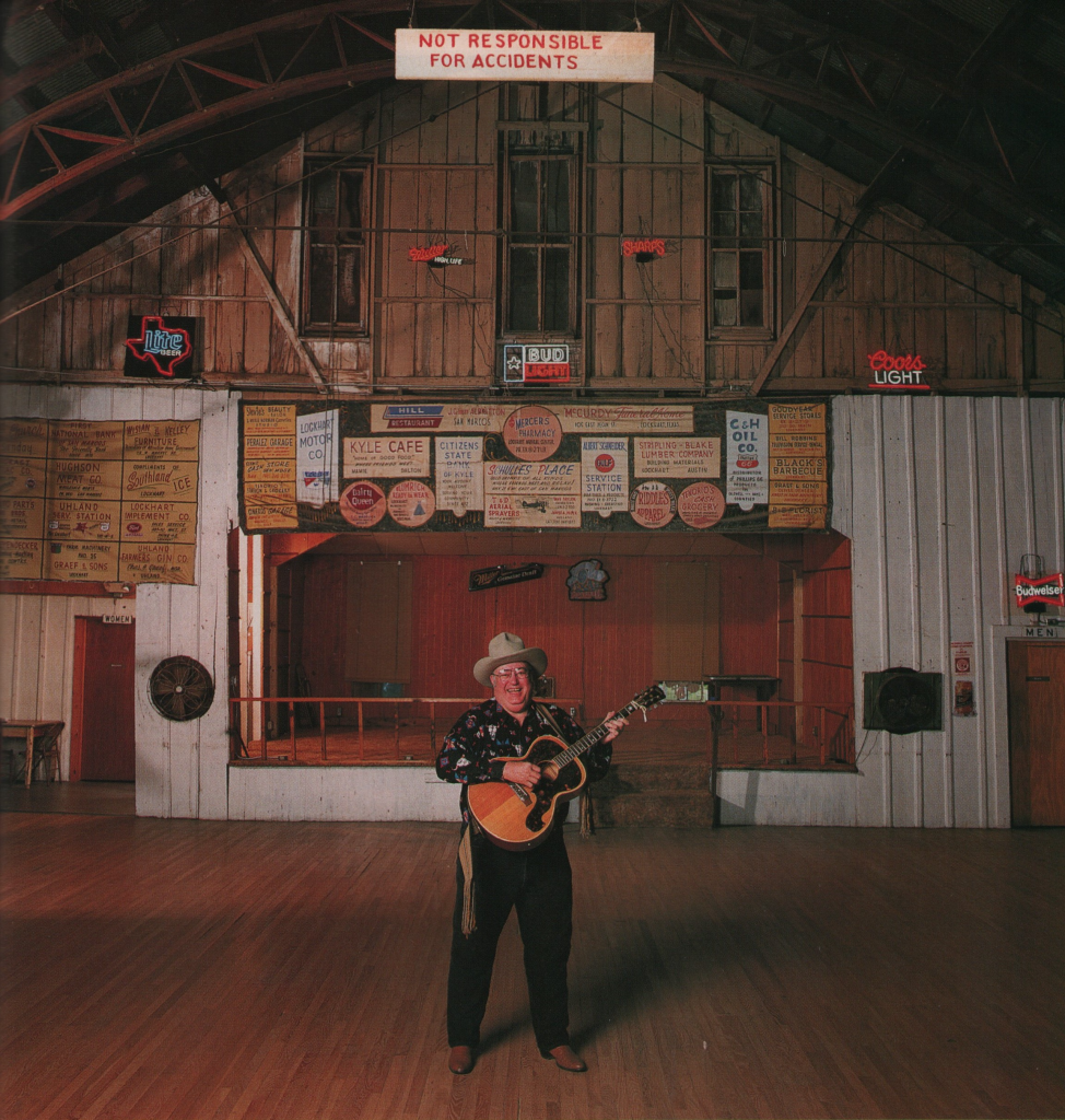 Don Walser standing with a guitar in the middle of a large wooden dance hall at Club 21 in Uhland, Texas. He is wearing a cowboy hat, Western-style clothing, and boots.