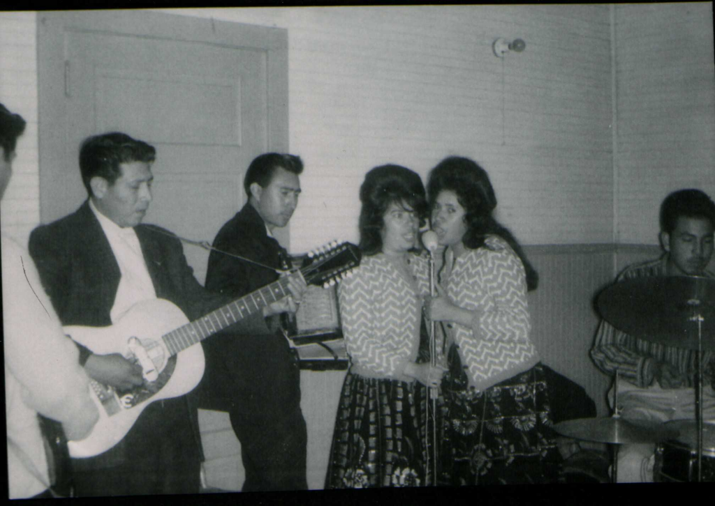 Two women, Petra Gonzalez and Mary Gonzalez, are singing into a shared microphone, wearing matching patterned tops and skirts. To their left, Jesse Gonzalez plays a guitar alongside another musician with a stringed instrument. A drummer is visible on the far right.