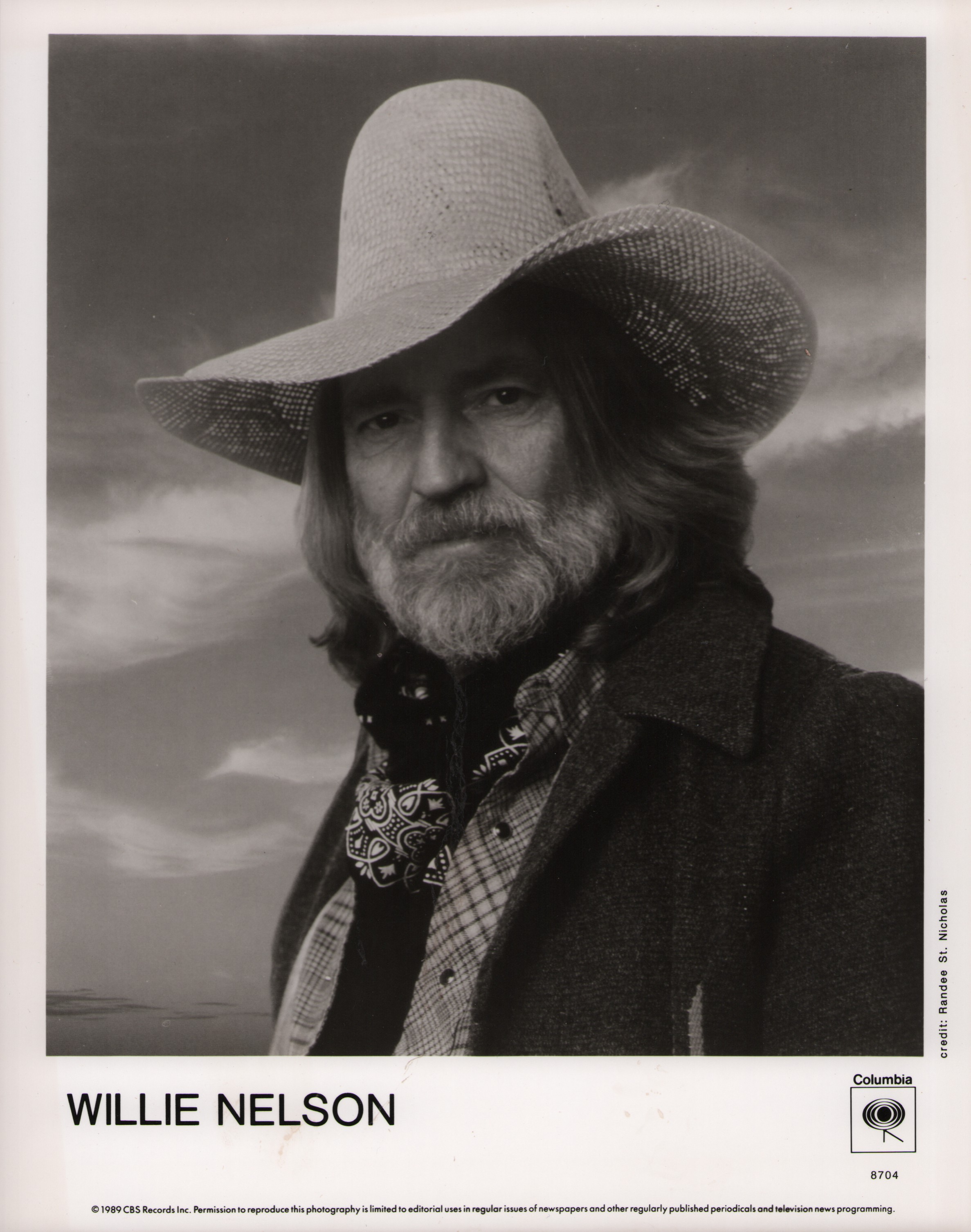 Willie Nelson wearing a cowboy hat, patterned bandana, and a coat over a plaid shirt.