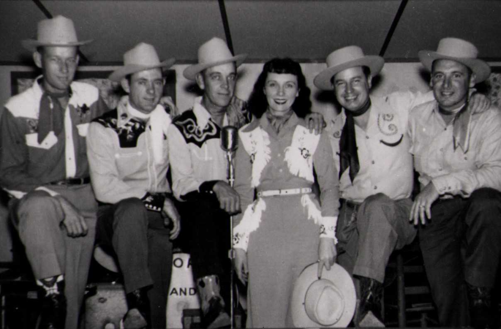 Dolores Farris and the Bluebonnet Boys, a group of five men and one woman dressed in Western-style outfits with cowboy hats. They are posed together with a microphone in front of them, smiling for the camera.