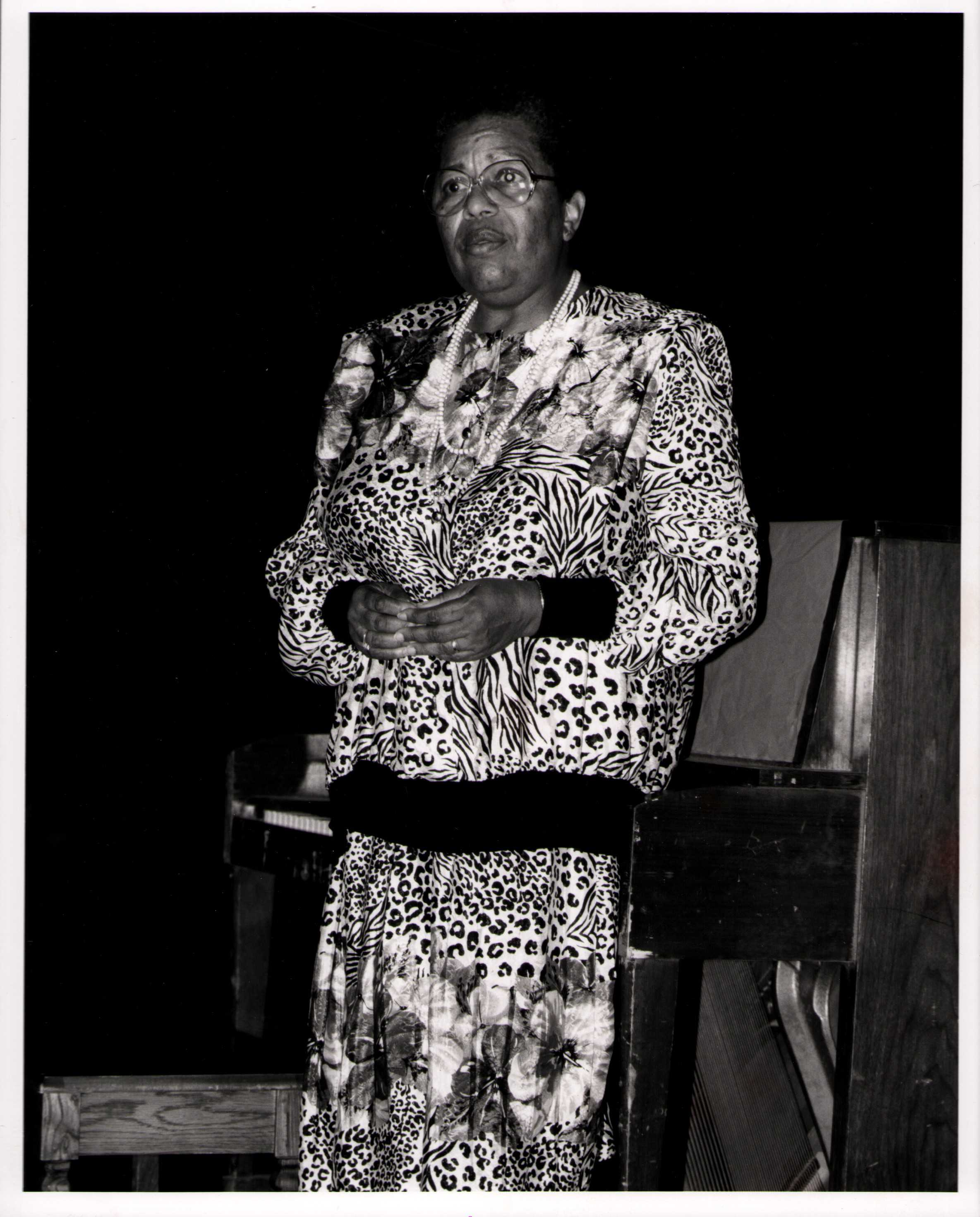 Dr. Beulah Agnes Curry Jones standing in front of a piano, wearing glasses and a patterned outfit featuring animal and floral prints. She is clasping her hands in front of her and looking slightly upward.