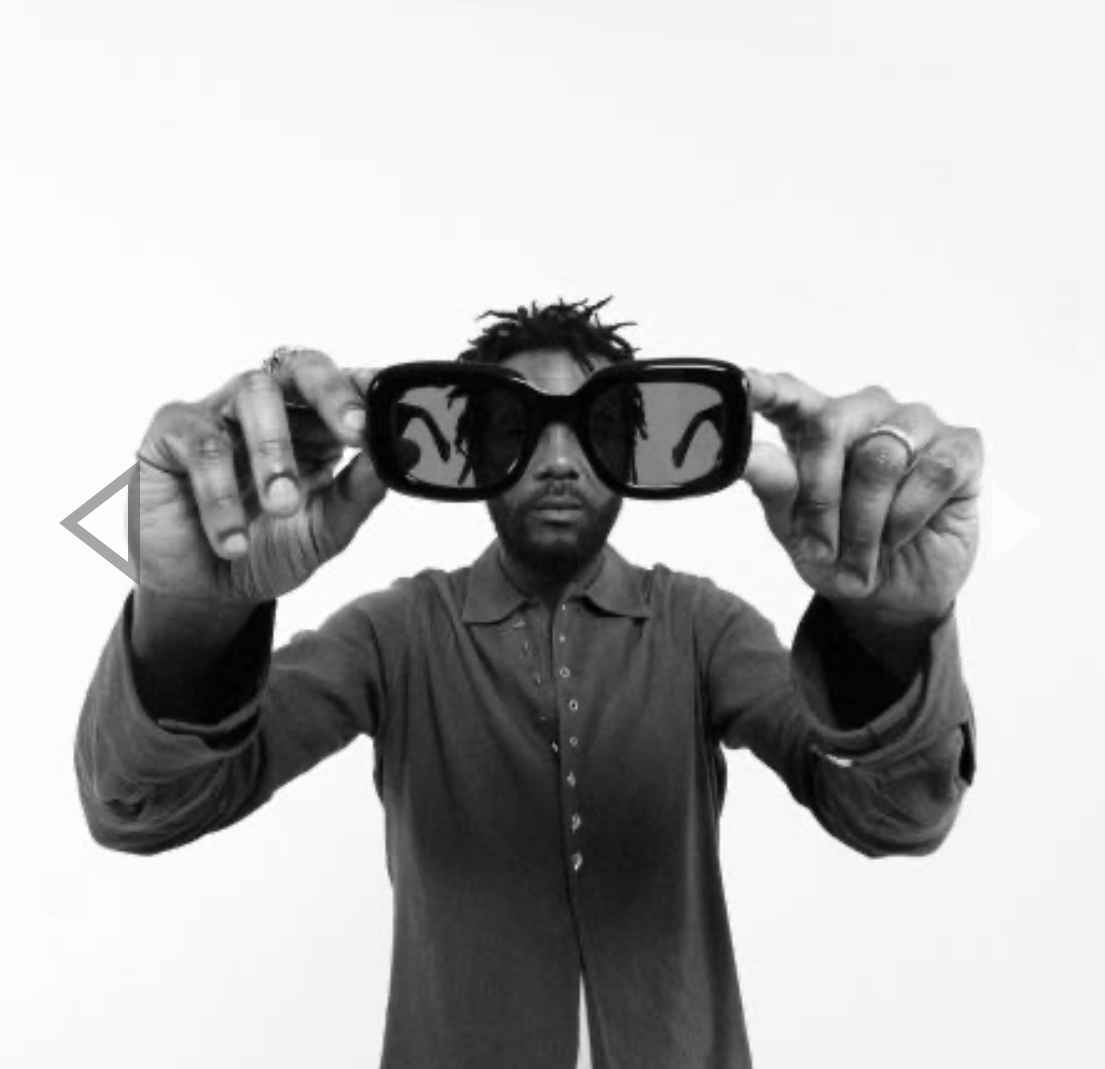 Gary Clark Jr. holding a pair of large sunglasses toward the camera, with his face visible through the lenses. He is wearing a button-up shirt and has a neutral expression against a plain white background.