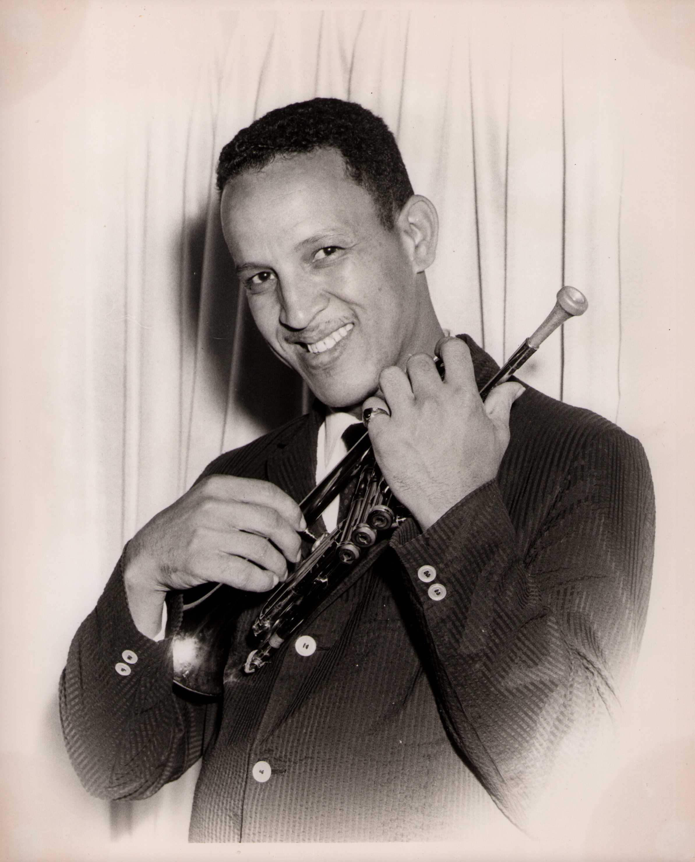 Gilbert Askey smiling while holding a trumpet. He is dressed in a pinstripe suit and tie, standing in front of a curtain backdrop.