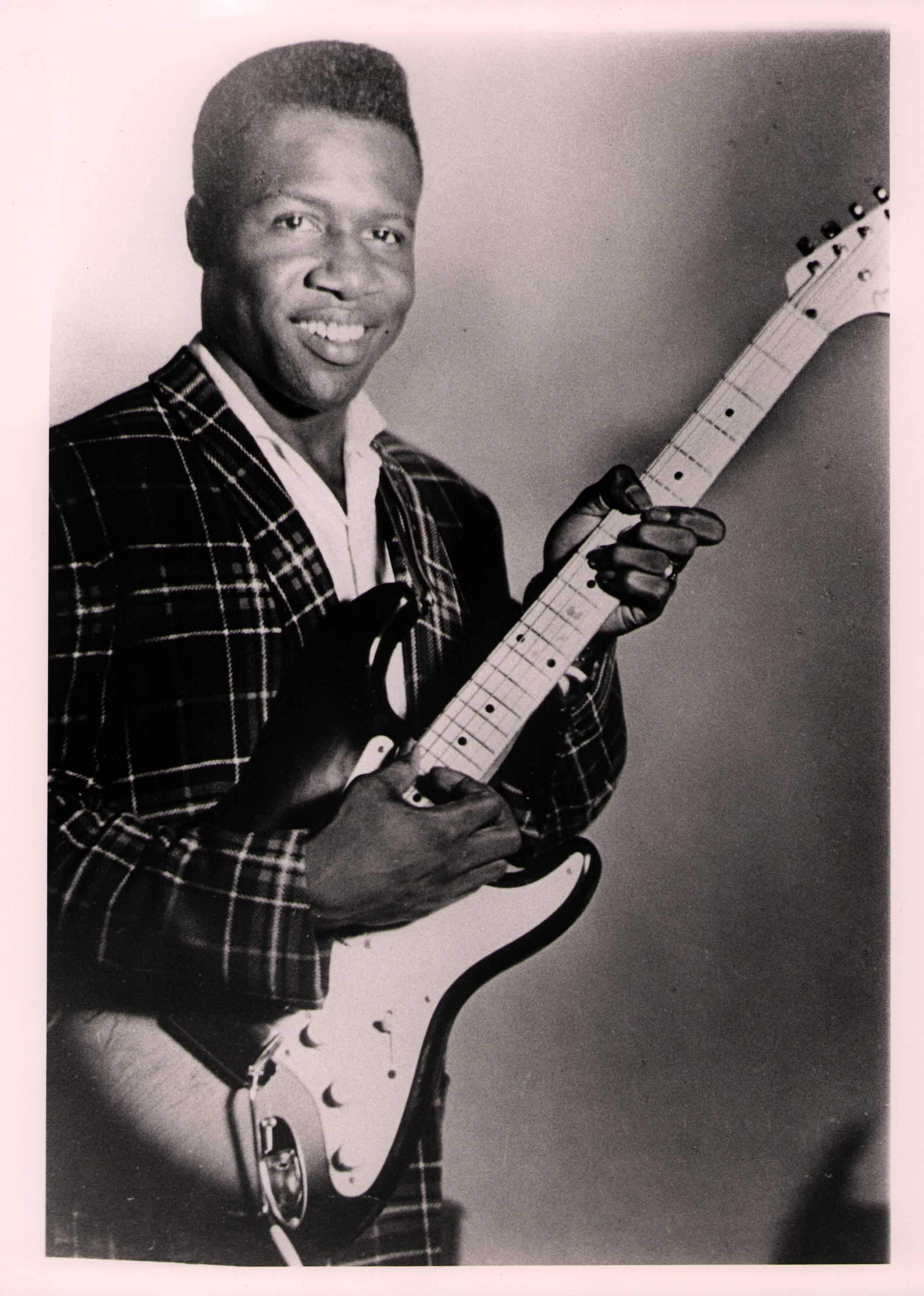 Henry 'Blues Boy' Hubbard smiling and holding an electric guitar. He is wearing a plaid suit jacket over a white shirt, standing against a plain background.