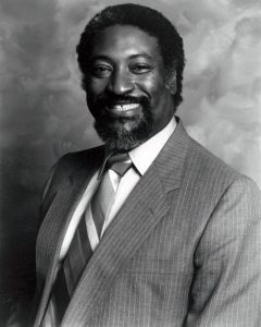 James Polk smiling, wearing a suit with a striped tie. He is posed against a softly textured backdrop, exuding a professional and approachable demeanor.