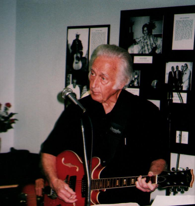 Jody Meredith playing an electric guitar during a performance, wearing a black shirt and standing in front of a microphone.