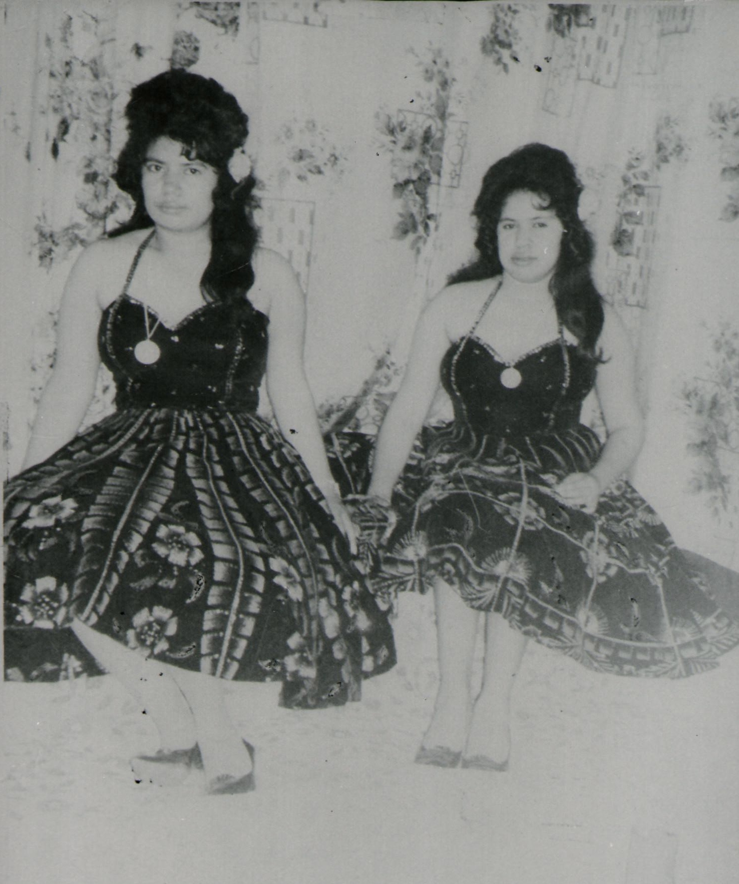 Las Hermanas Gonzalez, two women seated side by side, wearing matching patterned dresses with halter tops. Both have styled hair adorned with flowers.