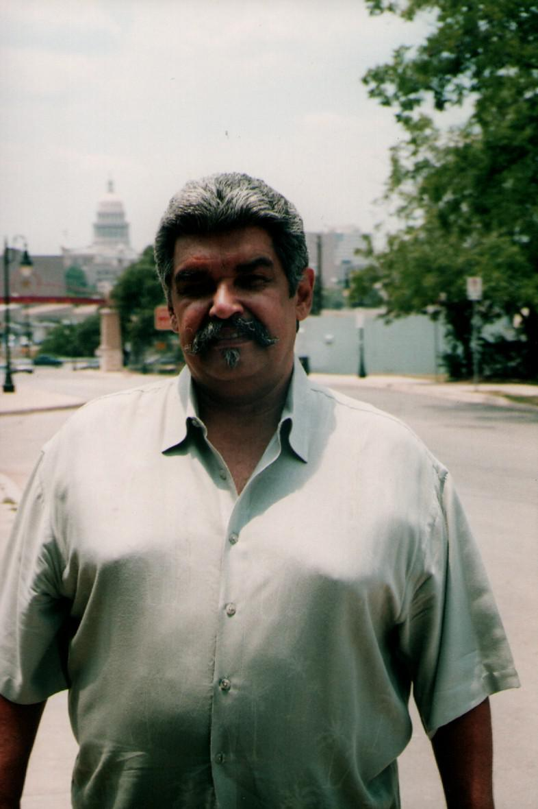 Leonard Davila, a man with a mustache and short gray hair, stands outdoors on a sunny day, wearing a light-colored button-up shirt.