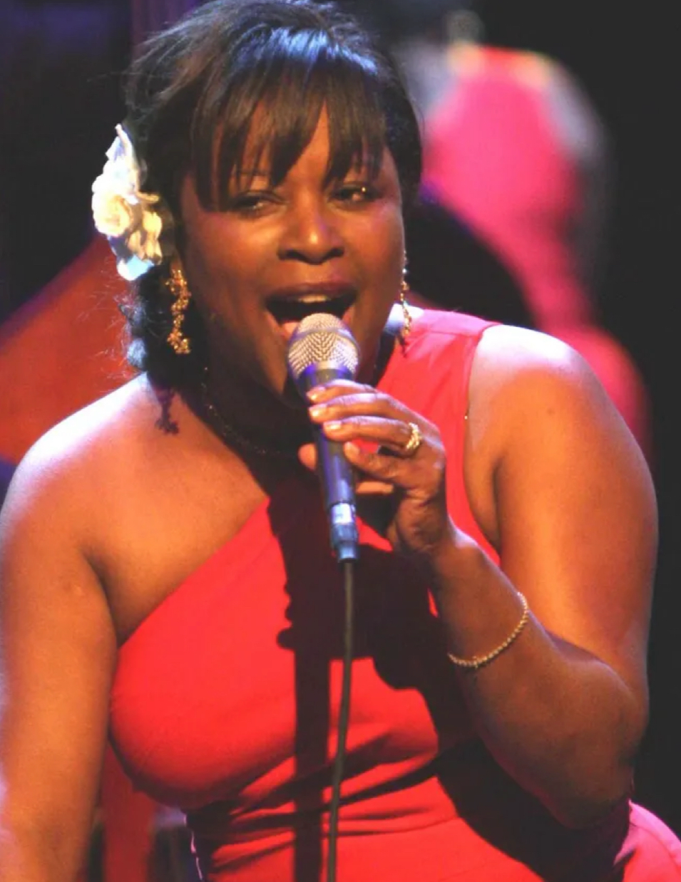 Pamela Hart performing on stage, holding a microphone and wearing a red one-shoulder dress. She has a white flower in her hair, gold jewelry, and is captured mid-song with a vibrant stage background.
