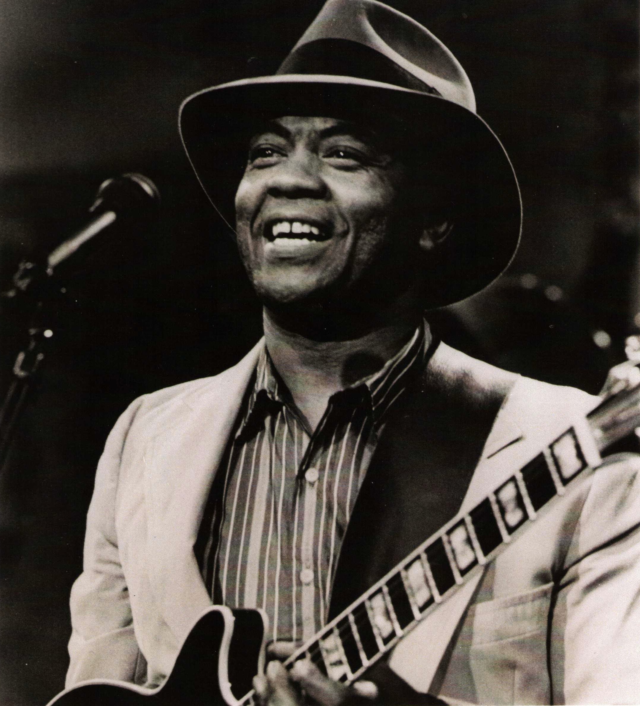 W.C. Clark smiling while performing on stage. He is wearing a wide-brimmed hat, a striped shirt, and a light-colored jacket, holding an electric guitar.