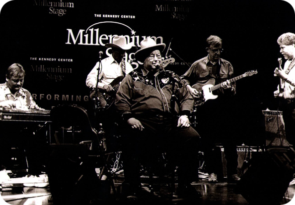 Don Walser performs with his band at the Kennedy Center, sitting in the spotlight while surrounded by band members playing guitars and other instruments.