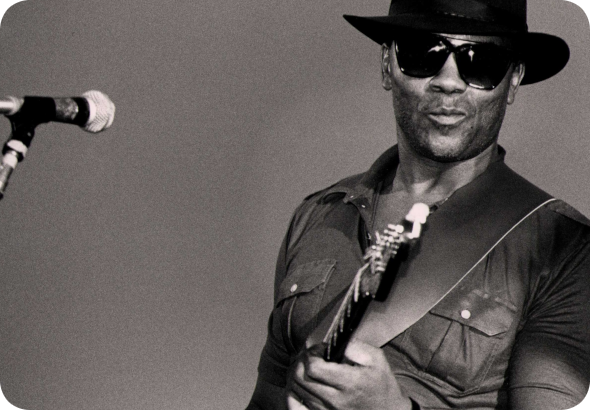 Black-and-white photo of W.C. Clark performing on stage. He is wearing sunglasses, a wide-brimmed hat, and a short-sleeve shirt, while playing the guitar. A microphone is visible in the foreground.