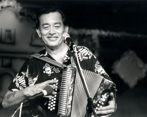 Flaco Jimenez playing the accordion and standing in front of a microphone. He is wearing a short-sleeve button-up patterned shirt.