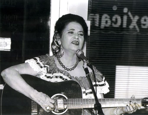Lydia Mendoza singing into a microphone while playing an acoustic guitar. She has dark, styled hair and is wearing a traditional blouse with lace detailing and a beaded necklace.