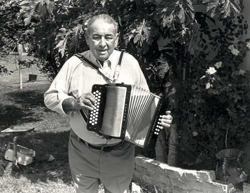 Narcisco Martinez standing outdoors playing an accordion. He has short, combed-back hair and is wearing a light-colored long-sleeve shirt and pants with suspenders.