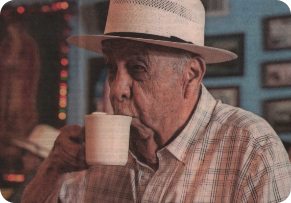 Color photograph of Johnny Degollado sipping coffee from a white mug at Joe's Bakery in Austin. He is wearing a light plaid shirt and a straw hat, with a blurred background featuring colorful lights and framed pictures.