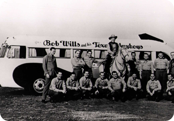 Black-and-white photograph of Bob Wills and his band, the Texas Playboys, taken in 1937. The group poses in front of their tour bus, which is labeled "Bob Wills and His Texas Playboys." Bob Wills sits on a white horse, wearing a Western-style outfit and cowboy hat. The band members, dressed in matching uniforms, are arranged in two rows, with some standing and others kneeling. 